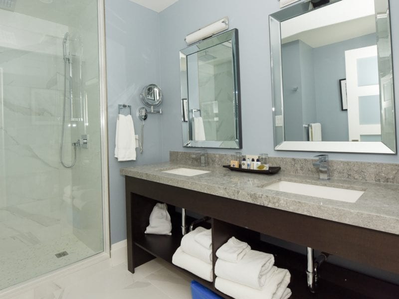 Modern bathroom with two mirrors above a double sink vanity. The countertop has toiletries and neatly folded towels underneath. Theres a glass-enclosed shower to the left with a towel hanging nearby, set against light blue walls. 124 on Queen Hotel and Spa | Hotel & Spa Niagara-on-the-lake | 1 (855) 988-4552