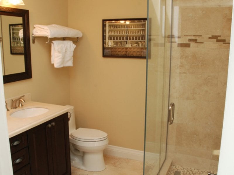 A bathroom with a modern, glass-enclosed shower. Theres a white toilet and a dark wood vanity with a sink and mirror. Towels are neatly stacked and hang on a rack above the toilet. A framed collection of miniature bottles decorates the wall. 124 on Queen Hotel and Spa | Hotel & Spa Niagara-on-the-lake | 1 (855) 988-4552