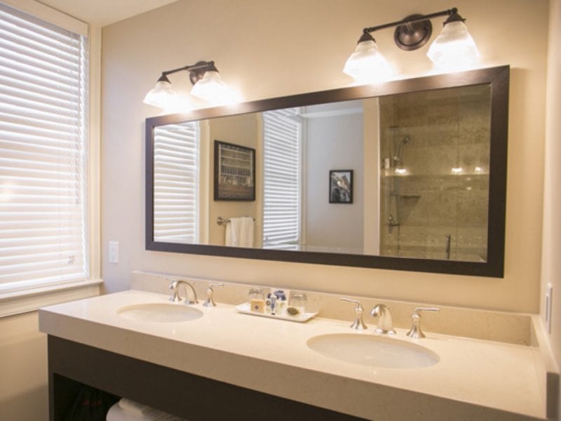 A modern bathroom features a double sink vanity with a large rectangular mirror above. Overhead, two wall-mounted lights illuminate the area. A window with blinds is on the left, and a glimpse of a shower area is visible in the mirrors reflection. 124 on Queen Hotel and Spa | Hotel & Spa Niagara-on-the-lake | 1 (855) 988-4552