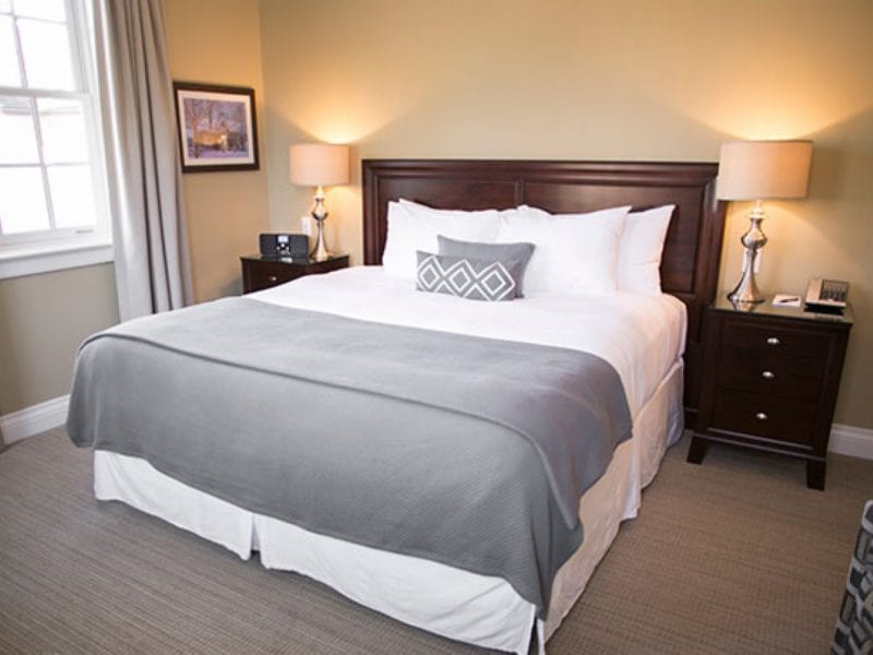 A cozy bedroom with a neatly made bed featuring white and gray bedding. Two bedside tables with matching lamps flank the bed. A painting hangs on the beige wall above one table, and a window with curtains is on the left. 124 on Queen Hotel and Spa | Hotel & Spa Niagara-on-the-lake | 1 (855) 988-4552