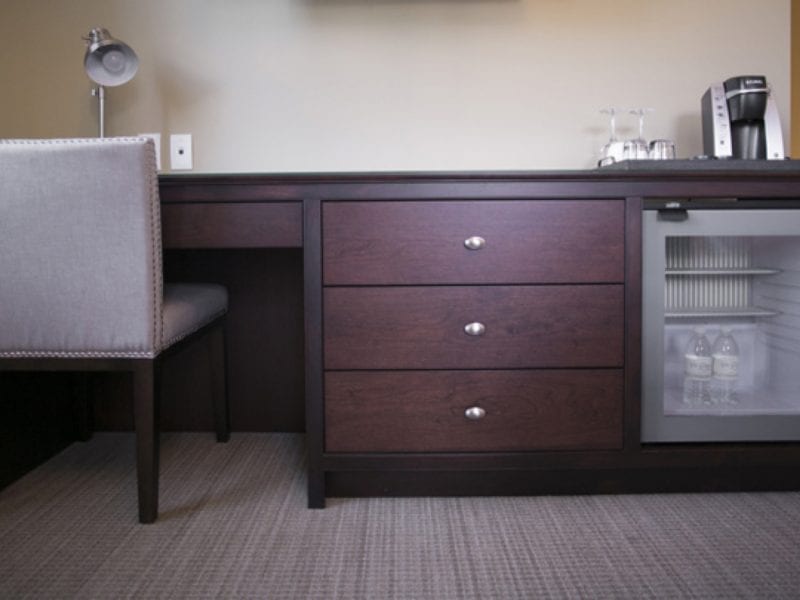 A hotel rooms desk with a lamp and a chair beside a mini-fridge containing bottled water. The desk has three drawers and a coffee maker with cups on the wooden surface. The room has a beige carpet and wall. 124 on Queen Hotel and Spa | Hotel & Spa Niagara-on-the-lake | 1 (855) 988-4552