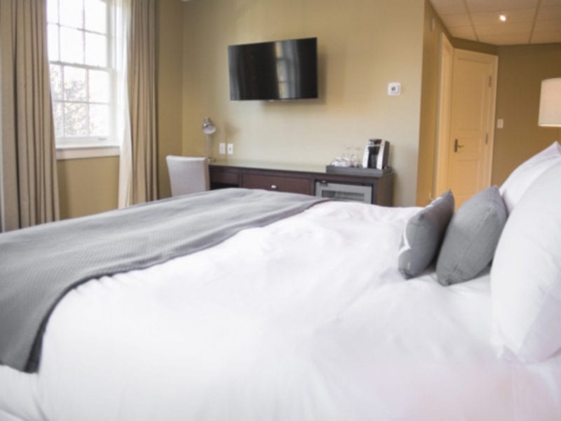 A cozy hotel room features a large bed with white linens and gray pillows. A wall-mounted TV and a mini-fridge are visible against a neutral-colored wall. A window with curtains allows natural light to brighten the space. 124 on Queen Hotel and Spa | Hotel & Spa Niagara-on-the-lake | 1 (855) 988-4552