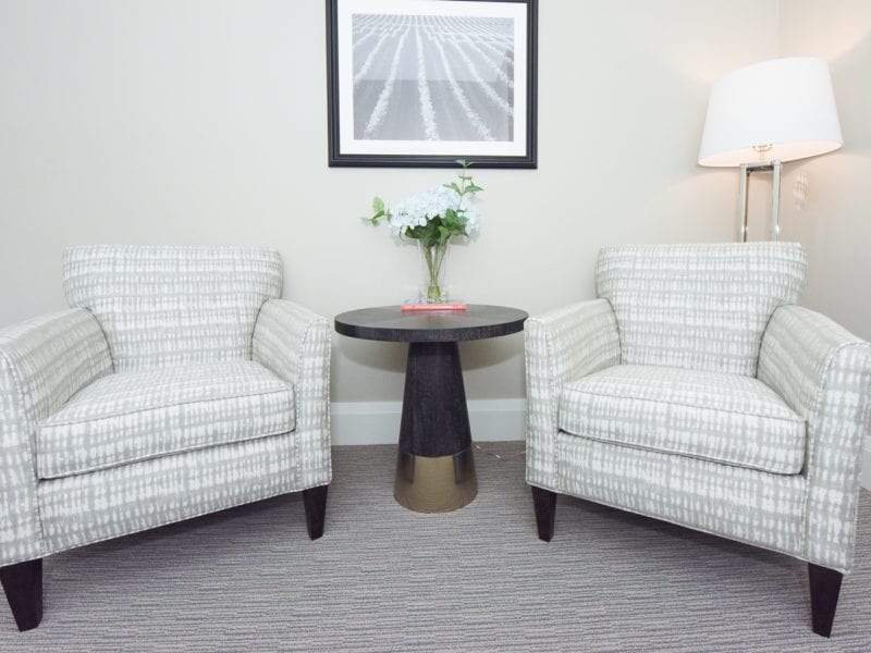 Two matching upholstered armchairs with a textured gray pattern are positioned on either side of a dark round table. A vase of white flowers and a book sit on the table. A floor lamp and a framed picture are on the light-colored wall. 124 on Queen Hotel and Spa | Hotel & Spa Niagara-on-the-lake | 1 (855) 988-4552