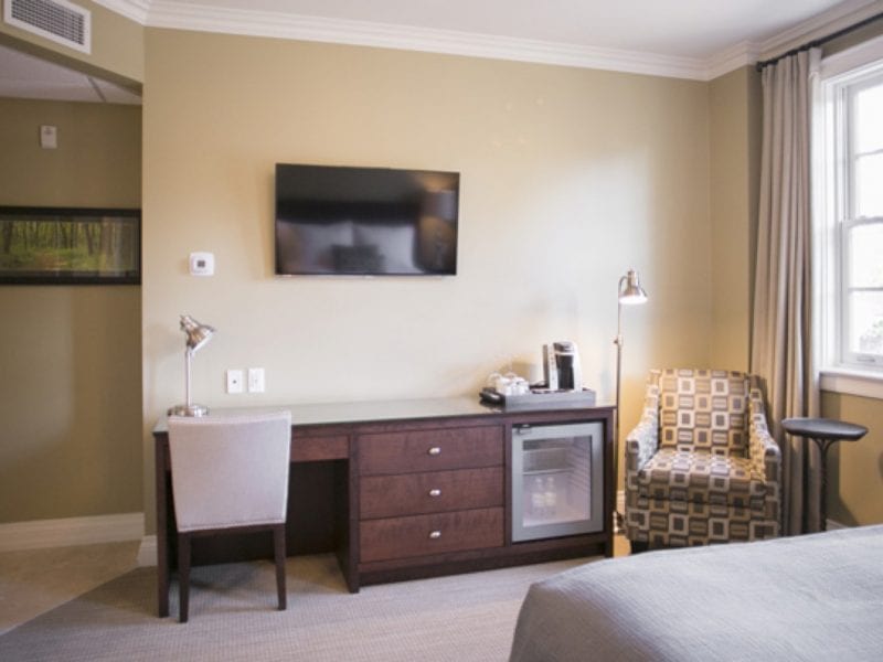 Hotel room interior with a flat-screen TV mounted on a beige wall above a dark wood desk. The desk has a coffee maker and lamp, flanked by a gray chair. A patterned armchair is beside a round table near a window with beige curtains. 124 on Queen Hotel and Spa | Hotel & Spa Niagara-on-the-lake | 1 (855) 988-4552