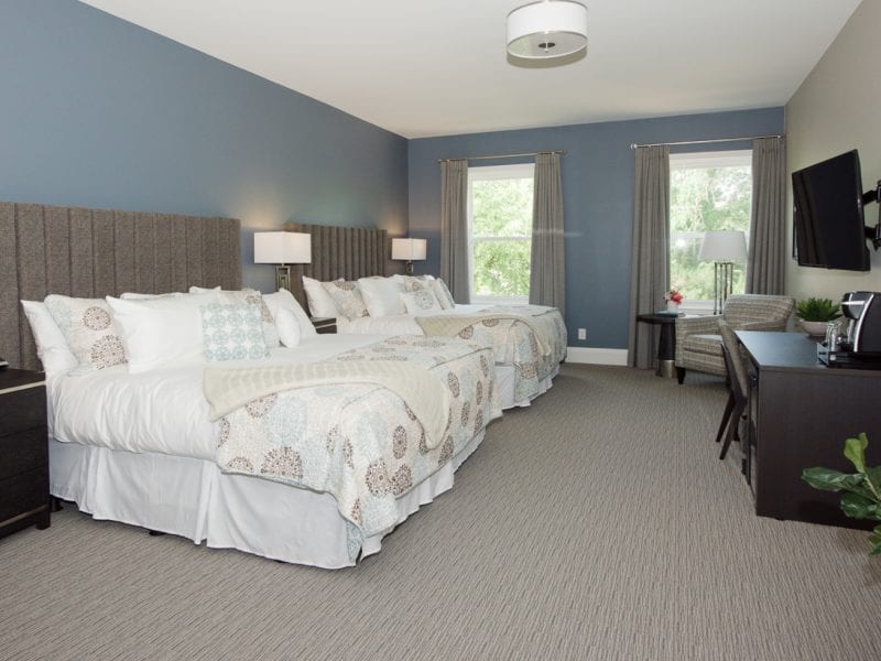 Hotel room with two large beds, gray headboards, and patterned pillows. A flat-screen TV is mounted on the wall opposite the beds. There’s a desk with a chair, a coffee maker, large windows with curtains, and a lounge chair with a small table. 124 on Queen Hotel and Spa | Hotel & Spa Niagara-on-the-lake | 1 (855) 988-4552