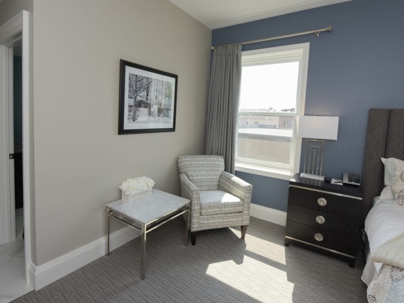 A modern bedroom with a blue accent wall and neutral decor. The room features a bed, nightstand with a lamp, and a gray armchair beside a small table with white flowers. A window lets in natural light, and a framed picture hangs on the wall. 124 on Queen Hotel and Spa | Hotel & Spa Niagara-on-the-lake | 1 (855) 988-4552