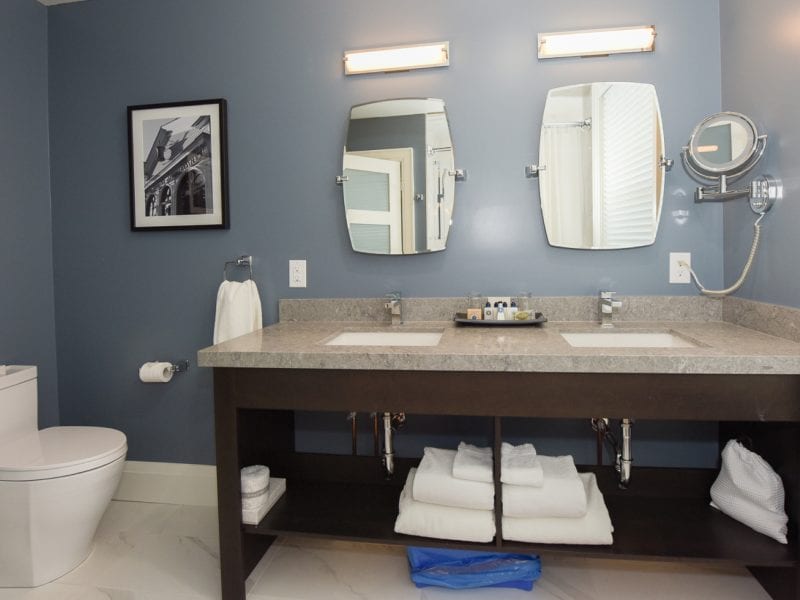 Modern bathroom with blue walls featuring a dual sink vanity with two mirrors and a light above each. A toilet is on the left, and a black-and-white photo hangs on the wall. Towels and toiletries are neatly arranged under the sinks. 124 on Queen Hotel and Spa | Hotel & Spa Niagara-on-the-lake | 1 (855) 988-4552