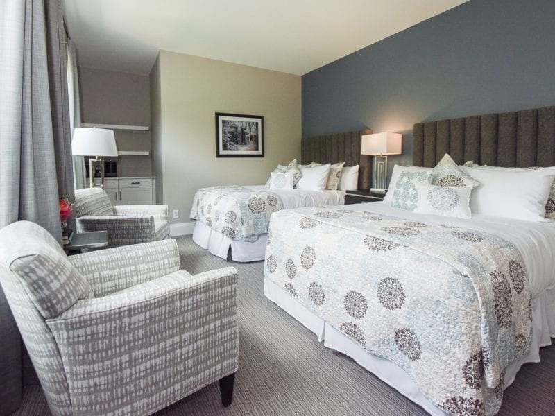 Hotel room with two queen beds, patterned bedding, and multiple pillows. The room has a soft gray color scheme, two armchairs by the window, a desk with a chair, and a framed picture on the wall. A modern lamp sits between the beds. 124 on Queen Hotel and Spa | Hotel & Spa Niagara-on-the-lake | 1 (855) 988-4552
