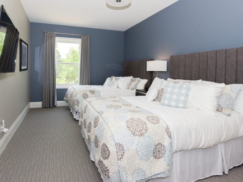 A bedroom with two neatly made beds, each covered in a light blue and beige patterned comforter. A large window with gray curtains allows natural light in. A wall-mounted TV is on the left, and a tall headboard runs along the wall behind the beds. 124 on Queen Hotel and Spa | Hotel & Spa Niagara-on-the-lake | 1 (855) 988-4552
