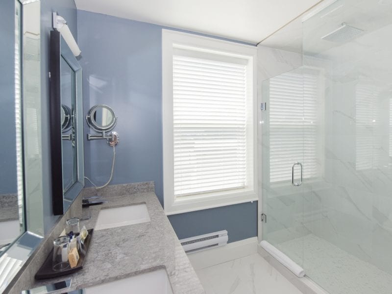 A modern bathroom with blue walls and marble countertops. It has two sinks, a mirrored cabinet, and a wall-mounted makeup mirror. A large window with blinds is next to a glass-enclosed shower with a rain showerhead. 124 on Queen Hotel and Spa | Hotel & Spa Niagara-on-the-lake | 1 (855) 988-4552
