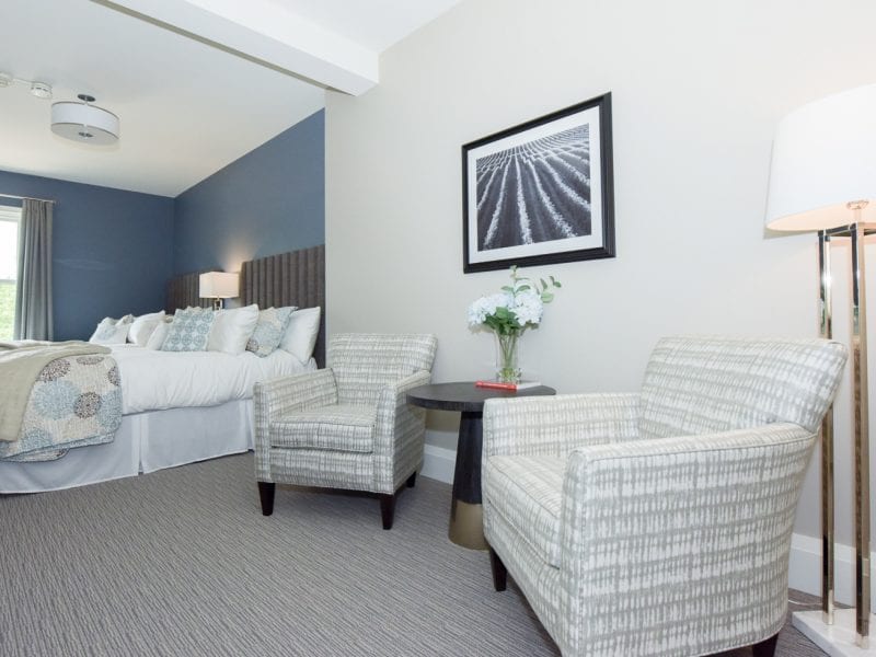 A modern hotel room features two patterned armchairs, a small round table with flowers, and a floor lamp. A neatly made bed with pillows and a throw blanket is visible. A framed black and white photo hangs on the wall. Large window with curtains. 124 on Queen Hotel and Spa | Hotel & Spa Niagara-on-the-lake | 1 (855) 988-4552