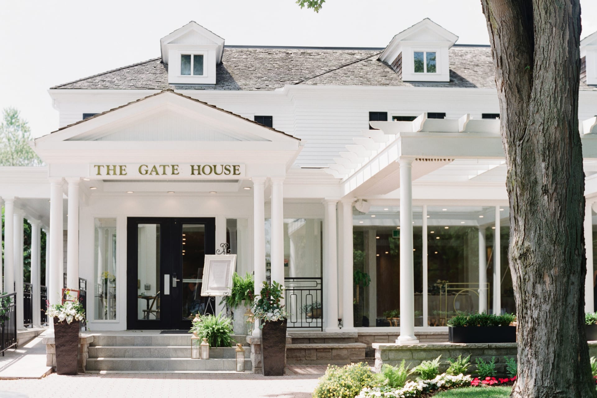 A white two-story building with the sign The Gate House above the entrance. The facade features large windows, a patio with white columns, and potted plants. A tree is in the foreground. 124 on Queen Hotel and Spa | Hotel & Spa Niagara-on-the-lake | 1 (855) 988-4552