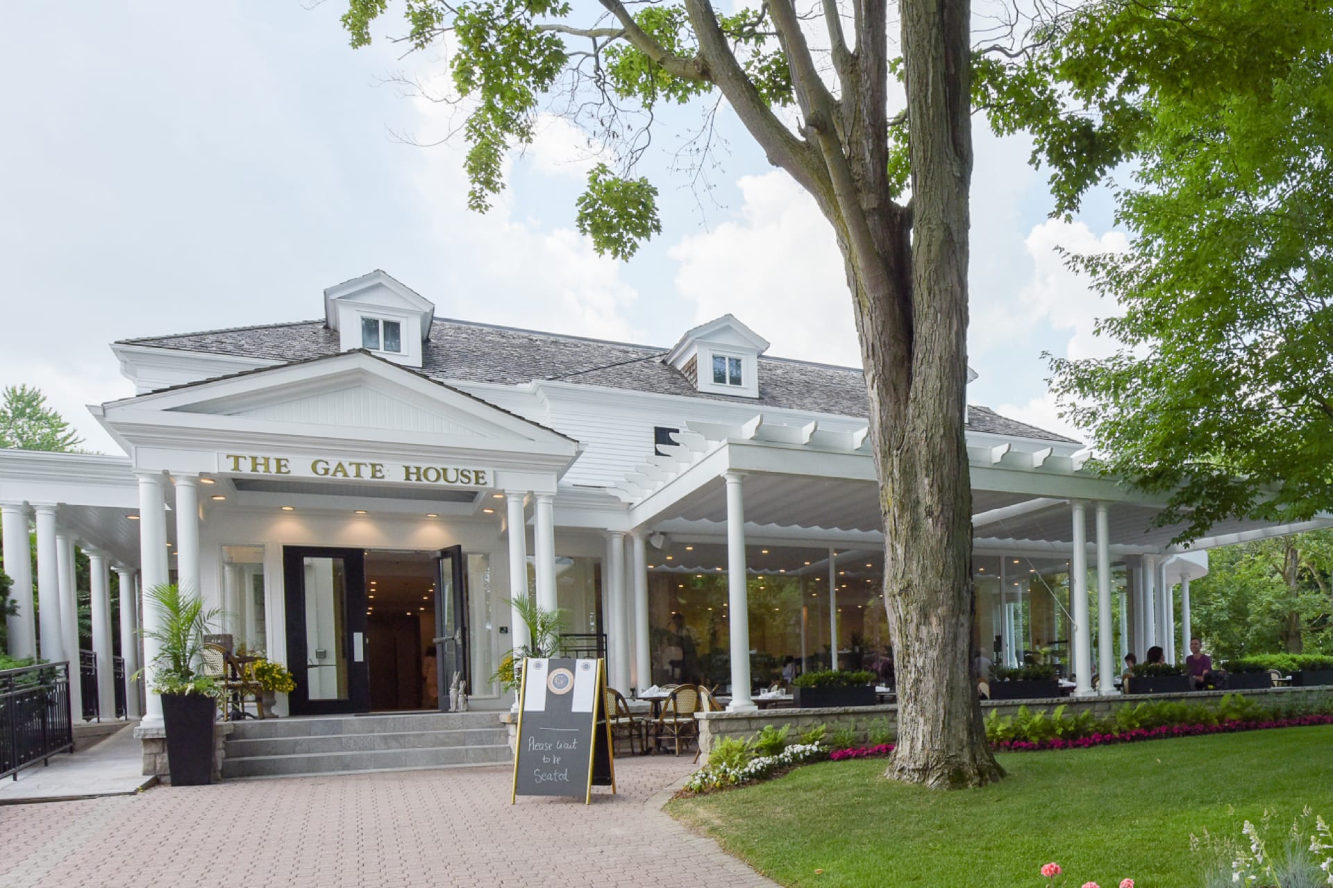 A white building with a sign The Gate House above the entrance. The structure features two small dormer windows and a covered patio with outdoor seating. A large tree stands in front, and a sign by the entrance advertises a special. 124 on Queen Hotel and Spa | Hotel & Spa Niagara-on-the-lake | 1 (855) 988-4552