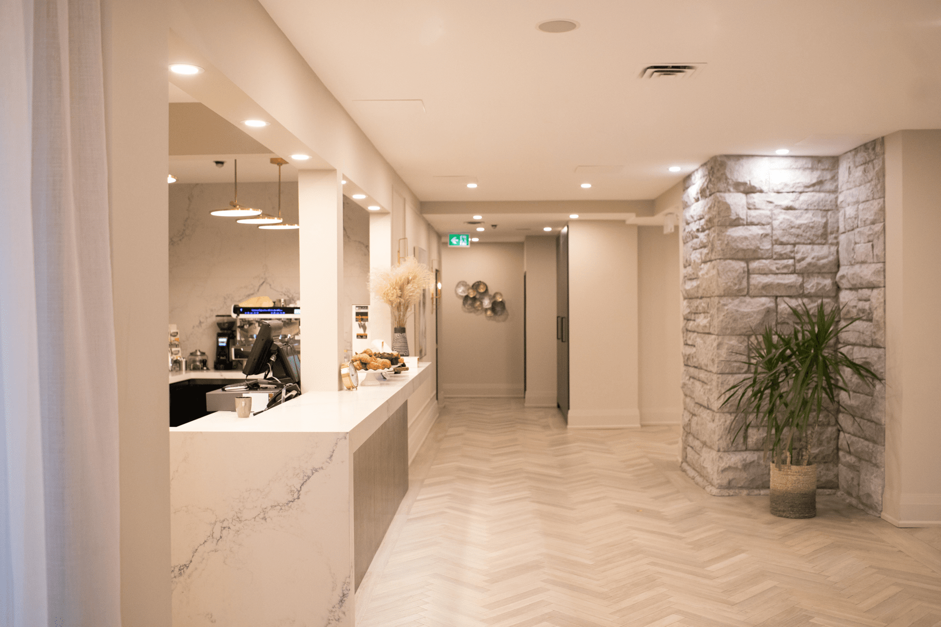 A modern, minimalist cafe interior with marble countertops and a wood-patterned floor. The cafe counter displays pastries under hanging lights. The room features a stone accent wall and a potted plant, with a doorway leading to another area. 124 on Queen Hotel and Spa | Hotel & Spa Niagara-on-the-lake | 1 (855) 988-4552