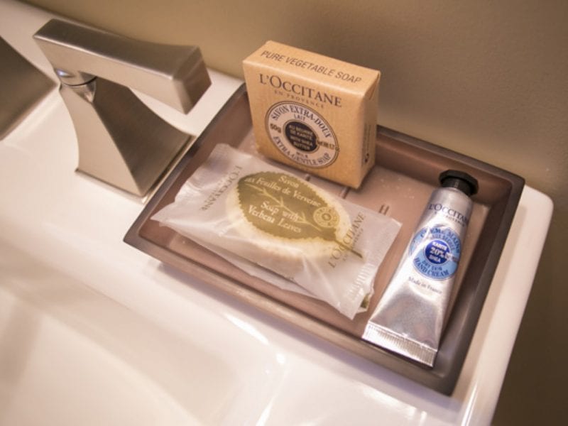 A bathroom countertop display with a modern faucet, featuring a bar of LOccitane soap, a small package of soap, and a LOccitane hand cream tube arranged on a rectangular tray. 124 on Queen Hotel and Spa | Hotel & Spa Niagara-on-the-lake | 1 (855) 988-4552