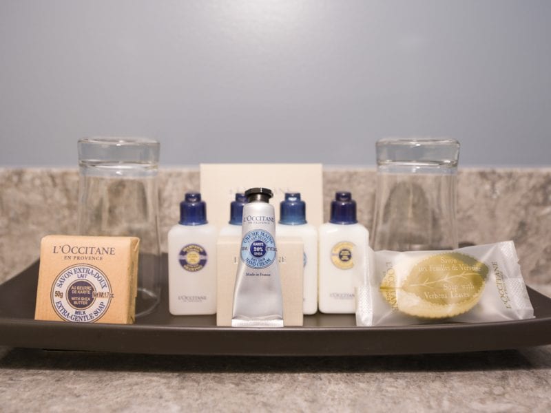 A tray on a marble countertop holds hotel toiletries, including soaps, lotion, and shampoo from LOccitane en Provence, alongside two upside-down glasses. The background is a light blue wall. 124 on Queen Hotel and Spa | Hotel & Spa Niagara-on-the-lake | 1 (855) 988-4552