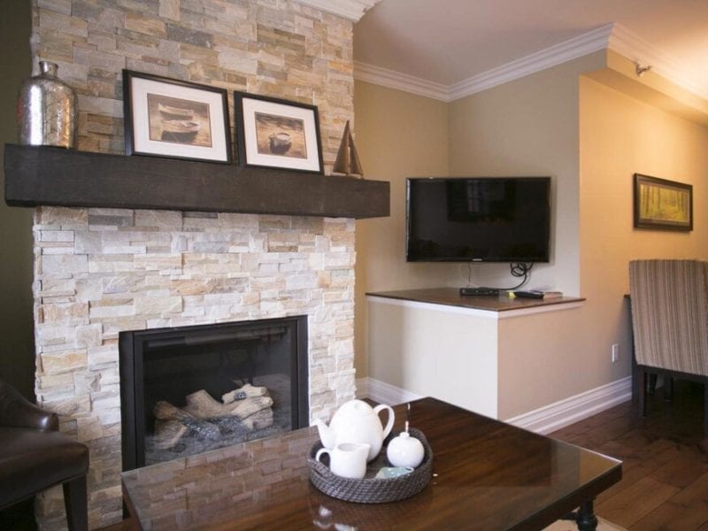 A cozy living room with a stone fireplace and a dark wooden mantel displaying framed art. A flat-screen TV is mounted on the beige wall nearby. A wooden coffee table holds a tray with a teapot and cups. The dining area is visible in the background. 124 on Queen Hotel and Spa | Hotel & Spa Niagara-on-the-lake | 1 (855) 988-4552