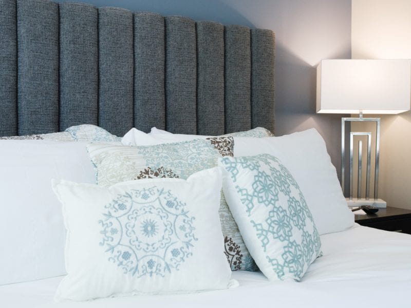 A neatly made bed with a gray padded headboard, featuring decorative pillows in white and light blue patterns. A modern silver table lamp sits on a bedside table to the right, casting a warm glow. 124 on Queen Hotel and Spa | Hotel & Spa Niagara-on-the-lake | 1 (855) 988-4552