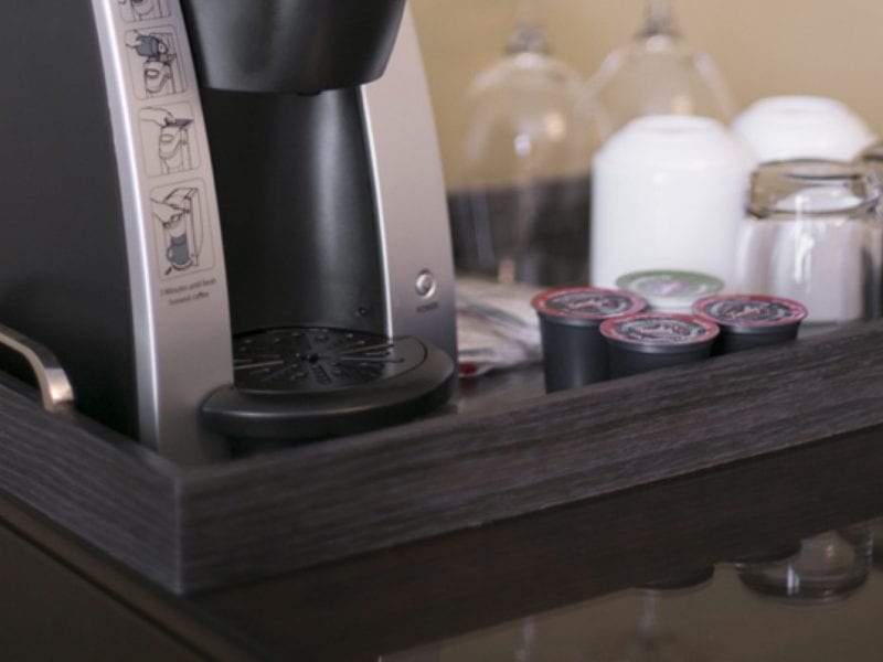 Close-up of a coffee maker on a dark wooden tray with several coffee pods beside it. In the background, there are upside-down glasses and white coffee mugs. 124 on Queen Hotel and Spa | Hotel & Spa Niagara-on-the-lake | 1 (855) 988-4552