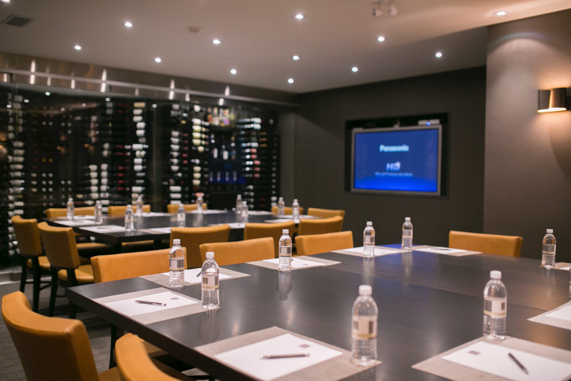 A conference room with a large table set with notepads, pens, and water bottles. There are several chairs around the table, and a large screen displays a presentation. A wall of wine bottles is visible in the background. 124 on Queen Hotel and Spa | Hotel & Spa Niagara-on-the-lake | 1 (855) 988-4552