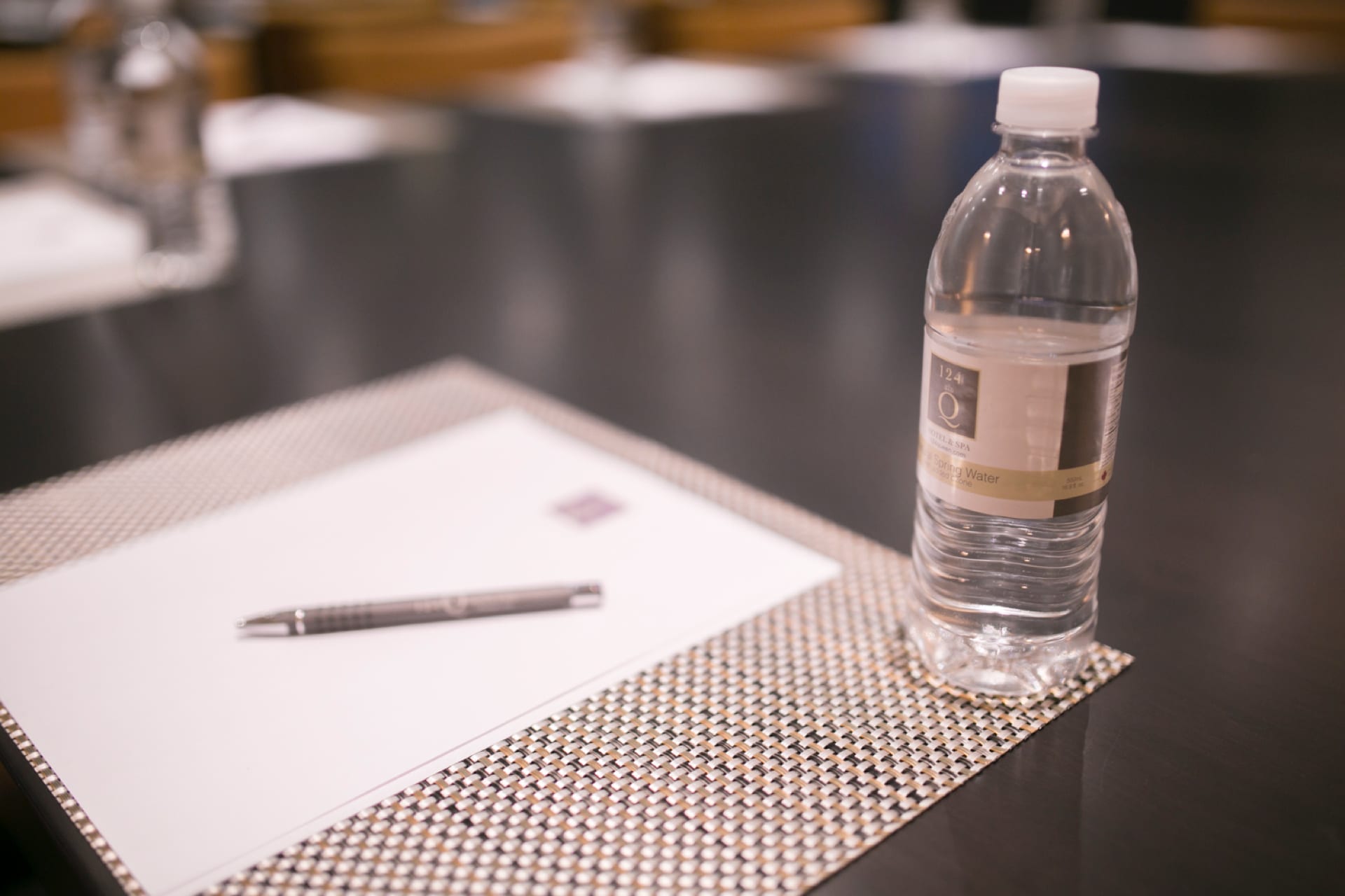 A conference table with a notepad and pen on a patterned mat, alongside a bottle of water. The focus is on the items, with more seats and similar setups visible in the blurred background. 124 on Queen Hotel and Spa | Hotel & Spa Niagara-on-the-lake | 1 (855) 988-4552