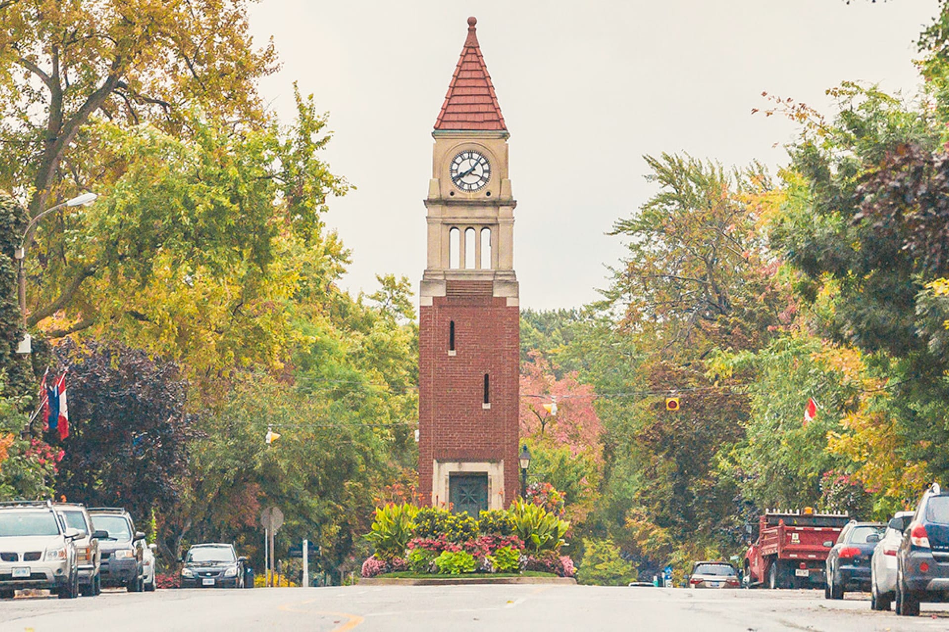 A tall brick clock tower with a red roof stands in the middle of a street lined with green and yellow trees. There are cars parked on both sides, and the sky is overcast. The scene captures an autumn setting. 124 on Queen Hotel and Spa | Hotel & Spa Niagara-on-the-lake | 1 (855) 988-4552