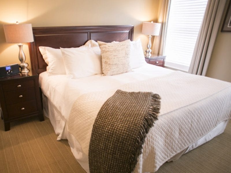 Cozy bedroom with a large bed featuring white bedding and a textured throw blanket. Two wooden nightstands, each with a lamp, flank the bed. A window with blinds and long curtains allows light into the room, which has beige walls and carpet. 124 on Queen Hotel and Spa | Hotel & Spa Niagara-on-the-lake | 1 (855) 988-4552