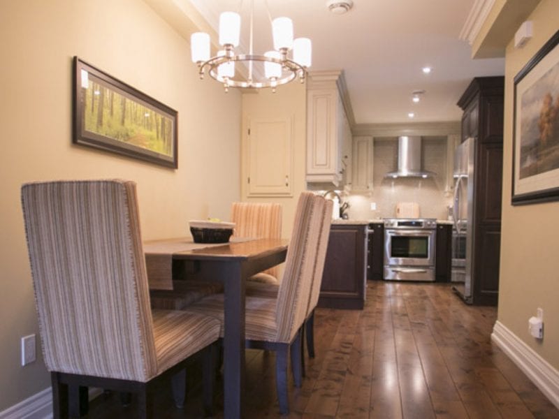 Dining area with a wooden table and six striped chairs. A modern kitchen is in the background with stainless steel appliances. The room has wooden floors, warm beige walls, a chandelier, and framed artwork. 124 on Queen Hotel and Spa | Hotel & Spa Niagara-on-the-lake | 1 (855) 988-4552
