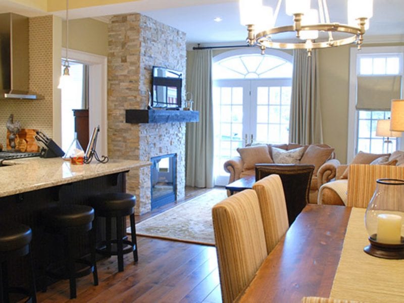 Open-plan living room and kitchen with a stone fireplace and wall-mounted TV. A wooden dining table, cushioned chairs, and a counter with bar stools are visible. French doors with curtains lead outside, and the floor is wooden. Neutral tones dominate. 124 on Queen Hotel and Spa | Hotel & Spa Niagara-on-the-lake | 1 (855) 988-4552