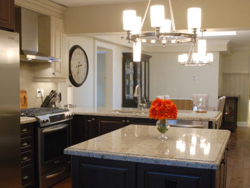 Modern kitchen with stainless steel appliances, including a refrigerator and oven. A large clock hangs on the wall. An island with a beige granite countertop features a vase of orange flowers, and a contemporary chandelier illuminates the space. 124 on Queen Hotel and Spa | Hotel & Spa Niagara-on-the-lake | 1 (855) 988-4552