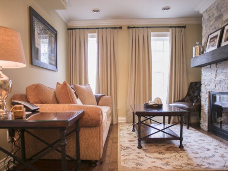 A cozy living room featuring a brown sofa with cushions, a dark wooden coffee table, and a stone fireplace. The room is adorned with framed art, a lamp on a side table, and cream-colored curtains covering two windows. A patterned rug lies under the table. 124 on Queen Hotel and Spa | Hotel & Spa Niagara-on-the-lake | 1 (855) 988-4552