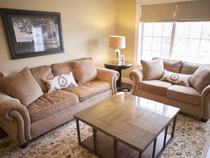 A cozy living room with a brown sofa and loveseat, both adorned with patterned cushions. A glass coffee table sits atop a decorative rug. A lamp and framed picture add warmth, while a large window lets in natural light. 124 on Queen Hotel and Spa | Hotel & Spa Niagara-on-the-lake | 1 (855) 988-4552