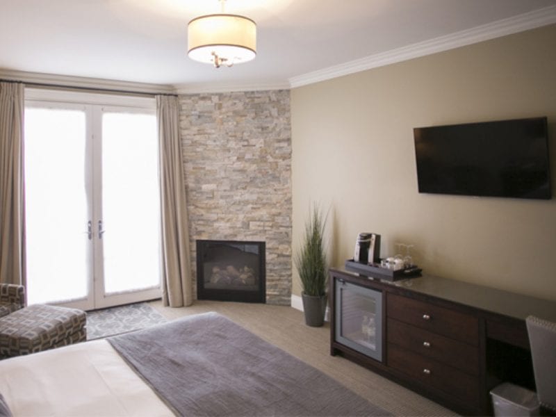 A cozy hotel room with a bed covered in white and gray linens. Theres a flat-screen TV on the wall, a stone fireplace, and a cabinet with a coffee maker. Large window doors are covered by curtains, and an armchair is in the corner. 124 on Queen Hotel and Spa | Hotel & Spa Niagara-on-the-lake | 1 (855) 988-4552