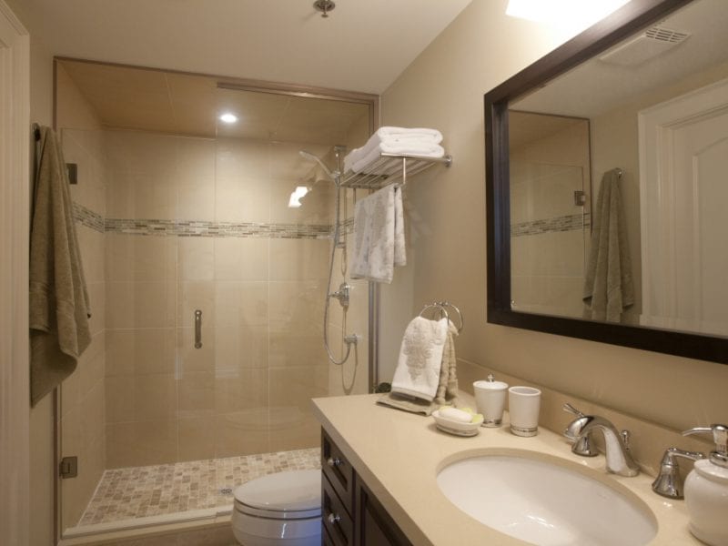 Modern bathroom with a glass-enclosed shower, beige tiles, and a wall-mounted showerhead. The vanity features a large mirror, sink, silver faucet, and toiletries. Towels hang on a rack, and the room is well-lit by overhead lighting. 124 on Queen Hotel and Spa | Hotel & Spa Niagara-on-the-lake | 1 (855) 988-4552