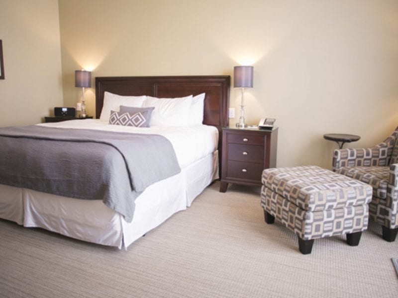 A hotel room featuring a large bed with white linens and a gray blanket. Two nightstands with lamps flank the bed. A patterned armchair and matching ottoman are in one corner, while a framed picture hangs on the wall. The carpet is light-colored. 124 on Queen Hotel and Spa | Hotel & Spa Niagara-on-the-lake | 1 (855) 988-4552