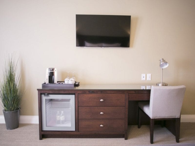 A hotel room desk setup featuring a minibar fridge, a wooden desk with drawers, a coffeemaker, an empty glass, and a silver desk lamp. A flat-screen TV is mounted on the wall above. A tall potted plant is to the left of the desk. 124 on Queen Hotel and Spa | Hotel & Spa Niagara-on-the-lake | 1 (855) 988-4552