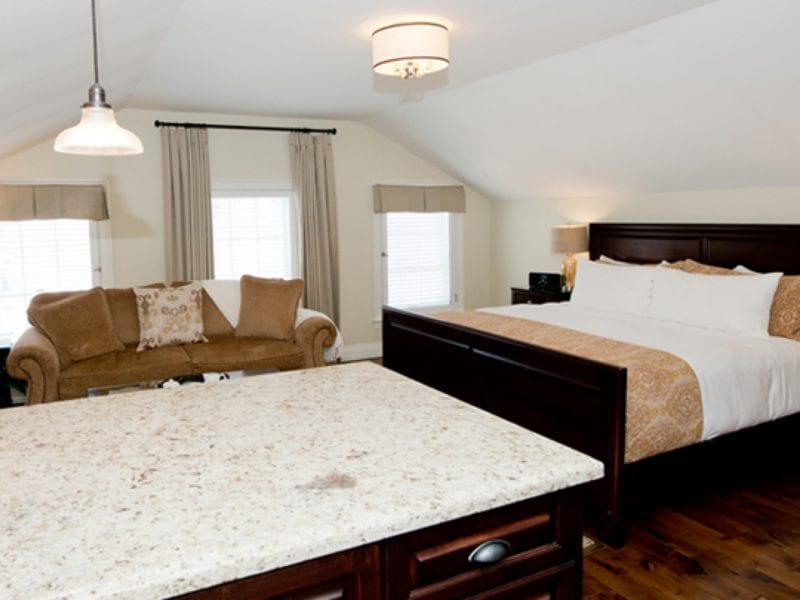 Bedroom with a dark wood bed, white and brown bedding, and a brown sofa. Granite countertop in the foreground and large windows with curtains and blinds. Ceiling lights illuminate the room. 124 on Queen Hotel and Spa | Hotel & Spa Niagara-on-the-lake | 1 (855) 988-4552