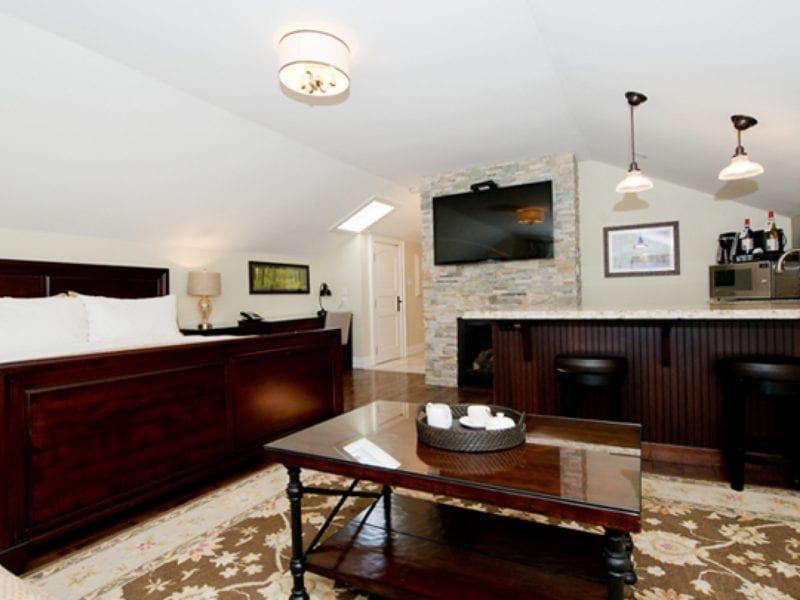 A cozy hotel room with a wooden bed, bedside tables, a coffee table, and a patterned rug. A stone accent wall holds a flat-screen TV. Theres a small bar area with barstools and hanging lights. Framed art decorates the walls. 124 on Queen Hotel and Spa | Hotel & Spa Niagara-on-the-lake | 1 (855) 988-4552