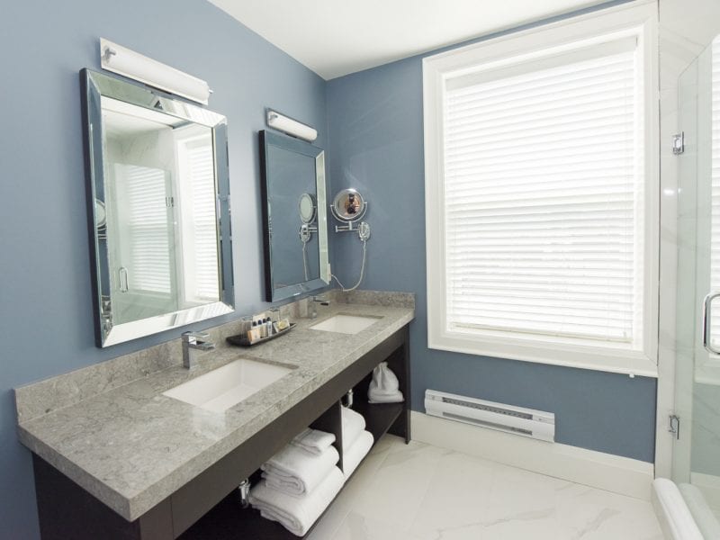 Modern bathroom with two sinks on a gray marble countertop, large mirrors above, and towels neatly stacked below. A large window with white blinds is on the right, next to a glass-enclosed shower with a chrome handle. Walls are painted in soft blue. 124 on Queen Hotel and Spa | Hotel & Spa Niagara-on-the-lake | 1 (855) 988-4552