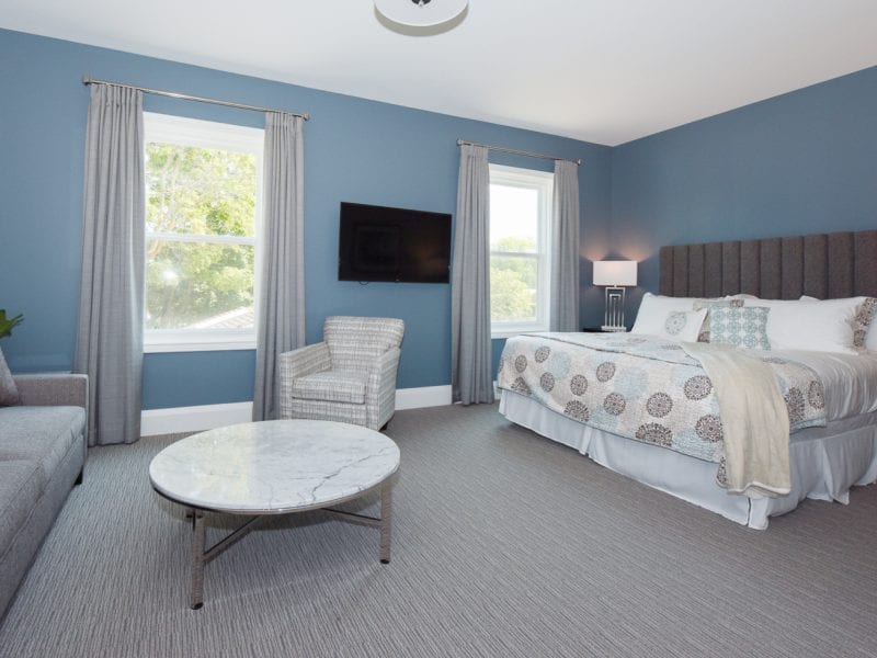 A bedroom with blue walls features a bed with patterned pillows, a gray upholstered headboard, and a light throw blanket. There’s a gray sofa, an armchair, a round marble coffee table, a wall-mounted TV, and two windows with gray curtains. 124 on Queen Hotel and Spa | Hotel & Spa Niagara-on-the-lake | 1 (855) 988-4552