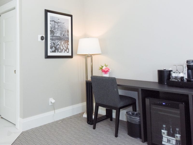 A minimalist hotel room corner with a black desk, a gray chair, and a lamp. Theres a coffee maker and ice bucket on the desk. A framed black-and-white photo hangs above. A mini-fridge and waste bin are below the desk. White sliding doors are visible. 124 on Queen Hotel and Spa | Hotel & Spa Niagara-on-the-lake | 1 (855) 988-4552