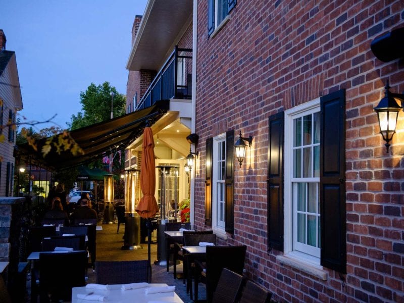 Outdoor dining area at dusk with tables and chairs along a brick building. Warm lighting from wall lanterns and orange umbrellas create a cozy atmosphere. Awnings extend over the seating area, with a visible tree and clear evening sky overhead. 124 on Queen Hotel and Spa | Hotel & Spa Niagara-on-the-lake | 1 (855) 988-4552