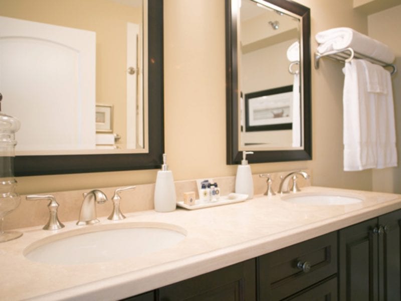 A bathroom with cream walls features a double sink vanity with two mirrors above it. The countertop holds soap dispensers and toiletries. White towels are neatly stacked on a rack to the right. The cabinet below is dark wood. 124 on Queen Hotel and Spa | Hotel & Spa Niagara-on-the-lake | 1 (855) 988-4552