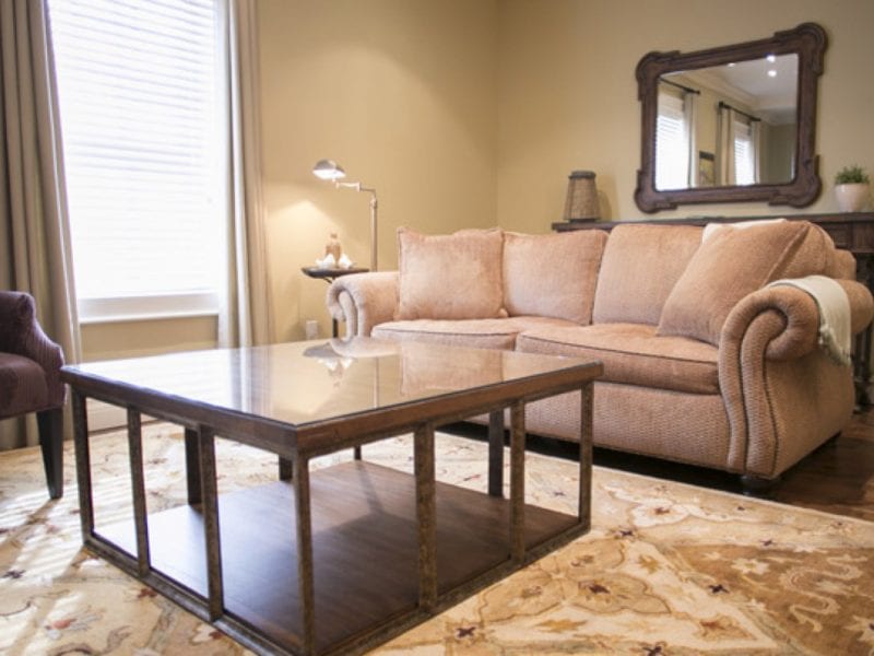 A cozy living room with a beige sofa, a glass-top coffee table, and an ornate mirror above a console table. The room features soft lighting, a patterned rug, and a window with closed blinds and curtains, creating a warm and inviting atmosphere. 124 on Queen Hotel and Spa | Hotel & Spa Niagara-on-the-lake | 1 (855) 988-4552