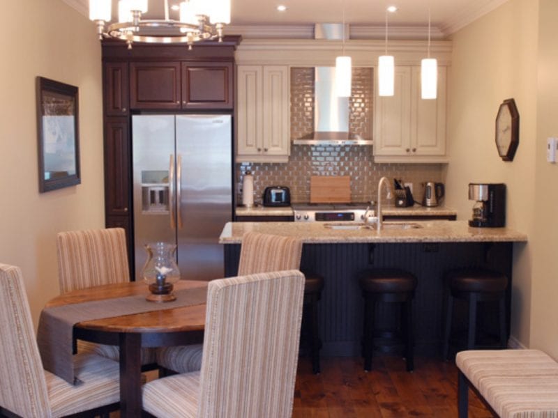 A cozy kitchen with a small dining area. The kitchen has dark cabinets, a metallic backsplash, a stainless steel fridge, and three pendant lights. The dining table has striped chairs, and a painting hangs on the beige wall. 124 on Queen Hotel and Spa | Hotel & Spa Niagara-on-the-lake | 1 (855) 988-4552
