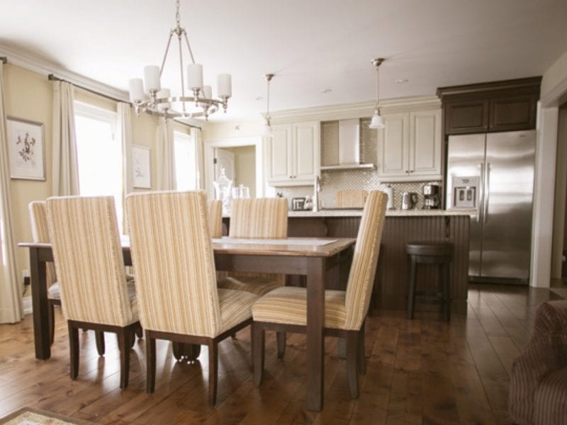 A cozy dining area with a wooden table and six striped upholstered chairs. A modern kitchen is visible in the background, featuring white cabinets, a stainless steel refrigerator, and hanging pendant lights. Warm lighting and wood flooring complete the scene. 124 on Queen Hotel and Spa | Hotel & Spa Niagara-on-the-lake | 1 (855) 988-4552