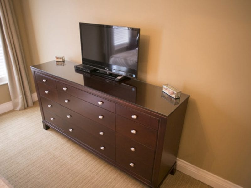 A dark wooden dresser with six drawers is topped with a flat-screen TV and two small boxes. Its placed against a beige wall, and a window with blinds and curtains is partially visible to the left. 124 on Queen Hotel and Spa | Hotel & Spa Niagara-on-the-lake | 1 (855) 988-4552
