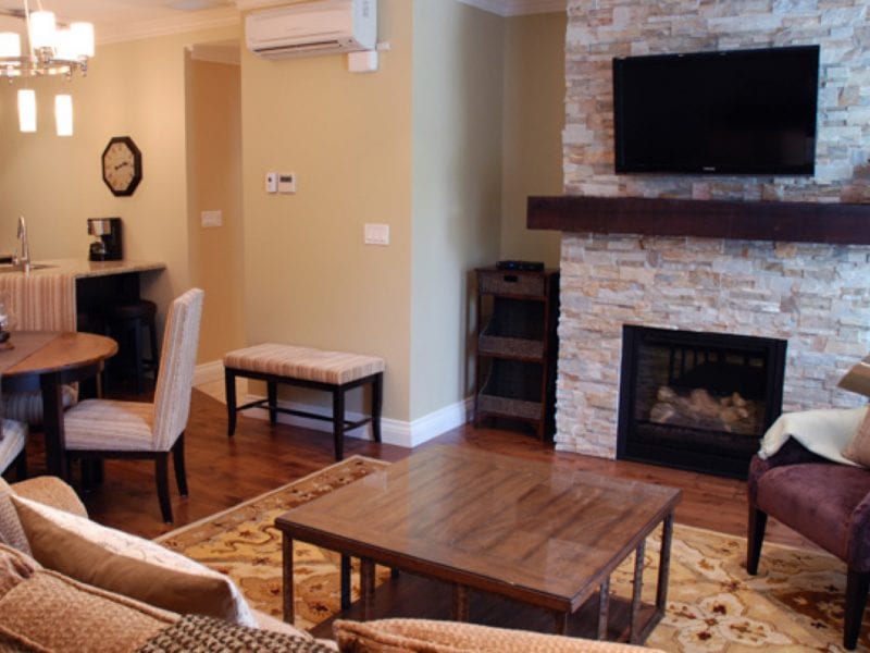 Cozy living room with a stone fireplace and wall-mounted TV. A wooden coffee table and beige sofa face the fireplace. A dining area with a round table and chairs is on the left. Warm lighting and earth-tone decor create a welcoming atmosphere. 124 on Queen Hotel and Spa | Hotel & Spa Niagara-on-the-lake | 1 (855) 988-4552