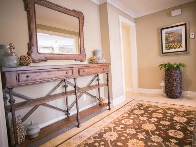 Elegant hallway with a wooden console table adorned with decorative vases and orbs. A large ornate mirror hangs above it. A patterned rug covers the floor, and a framed picture and plant are on the opposite wall. Neutral tones create a warm ambiance. 124 on Queen Hotel and Spa | Hotel & Spa Niagara-on-the-lake | 1 (855) 988-4552
