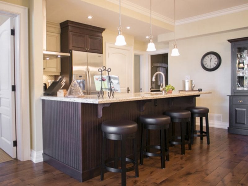 Modern kitchen with a wooden island, granite countertop, and four black bar stools. Hanging pendant lights illuminate the space. A clock and glass cabinet are visible in the background, with hardwood flooring throughout. 124 on Queen Hotel and Spa | Hotel & Spa Niagara-on-the-lake | 1 (855) 988-4552